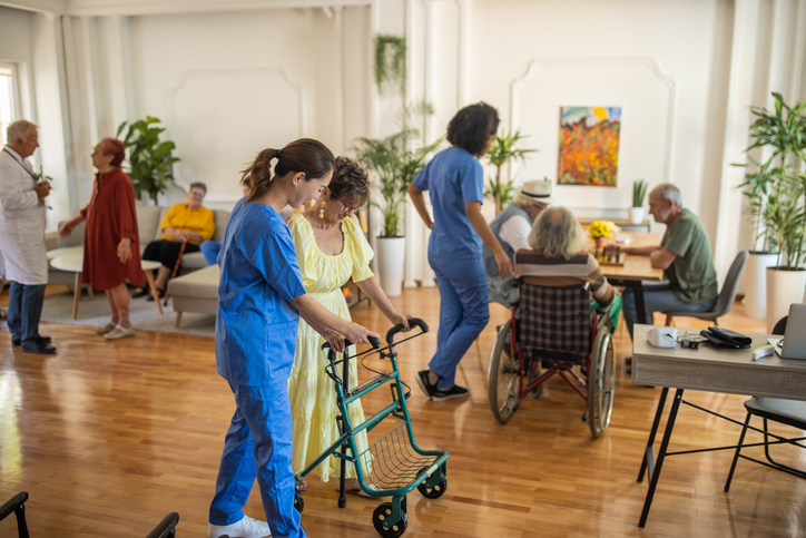 A nurse wearing blue scrubs assisting a lady wearing a yellow dress use a walker.