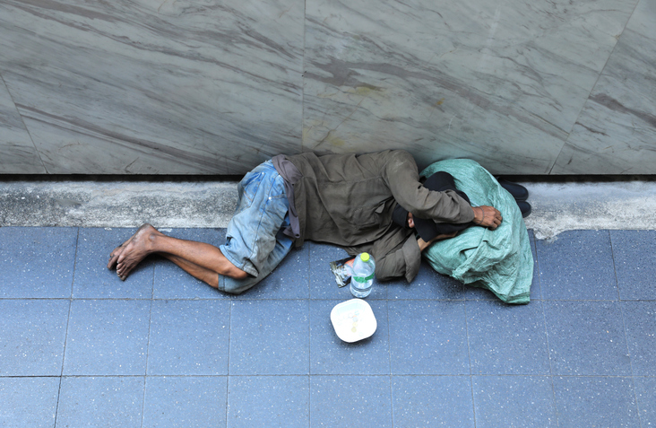 A man who appears dirty, laying on a sidewalk wearing ripped jeans and a dirty grey jacket has a blue tarp over his head while he sleeps.